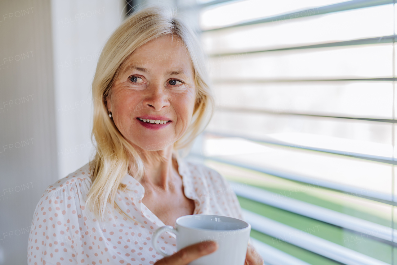 An attractive senior woman drinking tea and standing by window at home.