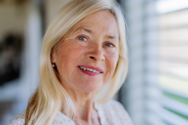 An attractive senior woman standing by window at home, looking at camera and smiling.