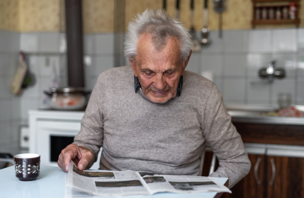 A portrait of elderly man sitting at the table indoors at home, reading newspapers.