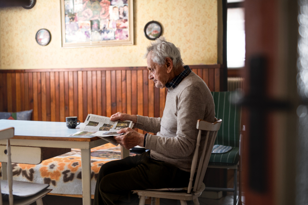 A portrait of elderly man sitting at the table indoors at home, reading newspapers.