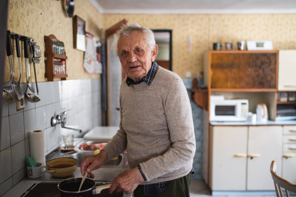 A portrait of elderly man cooking on stove indoors at home, stirring and looking at camera.