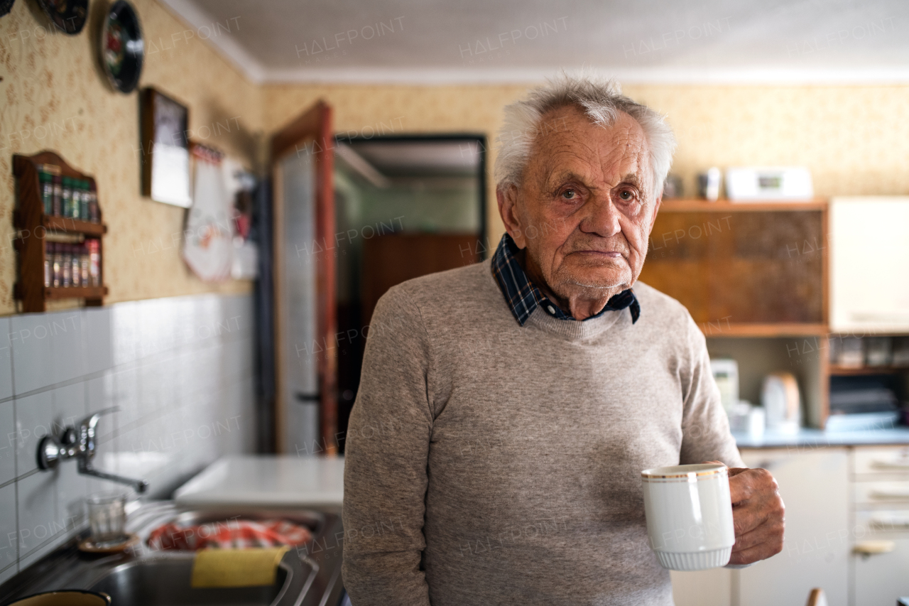 A portrait of elderly man with cup of tea standing indoors at home, looking at camera.