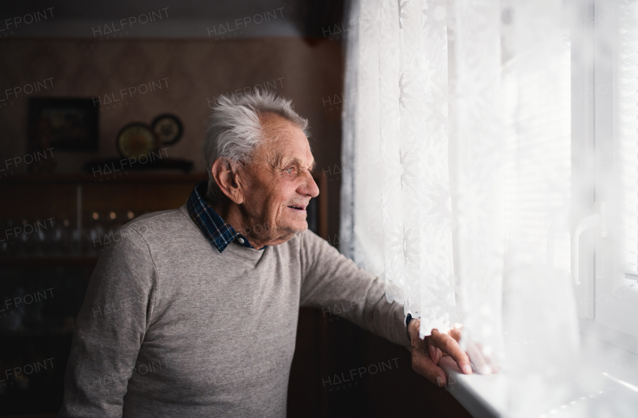 A portrait of elderly man standing indoors at home, looking out through window.