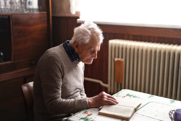 A portrait of elderly man sitting at the table indoors at home, reading book.