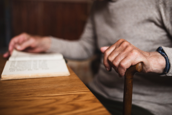 An unrecognizable elderly man sitting at the table indoors at home, reading book.