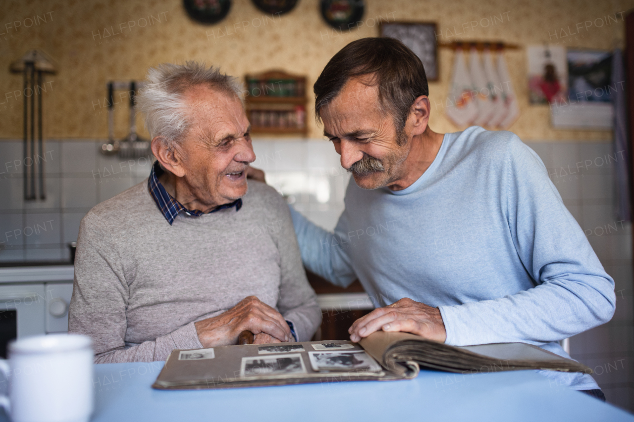 A portrait of man with elderly father sitting at the table indoors at home, looking at photo album.