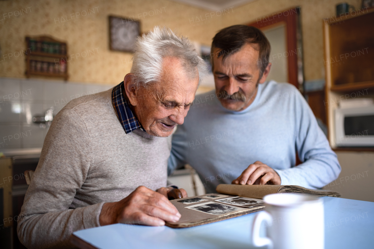 A portrait of man with elderly father sitting at the table indoors at home, looking at photo album.