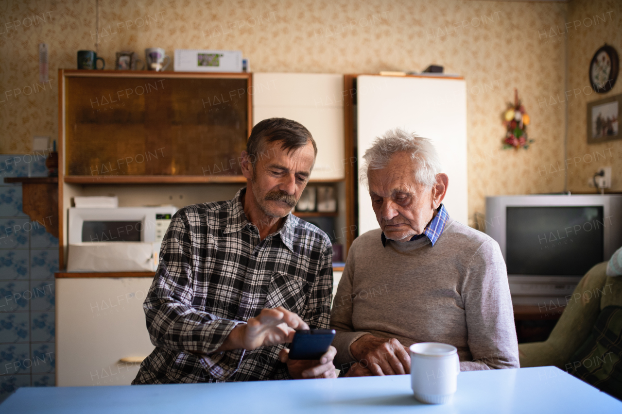 A portrait of man with elderly father sitting at the table indoors at home, using smartphone.