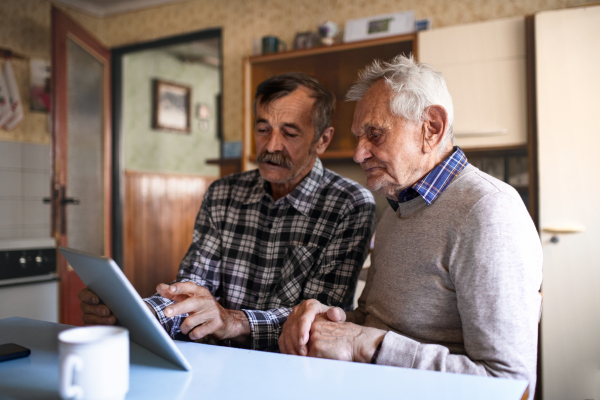 A portrait of man with elderly father sitting at the table indoors at home, using tablet.