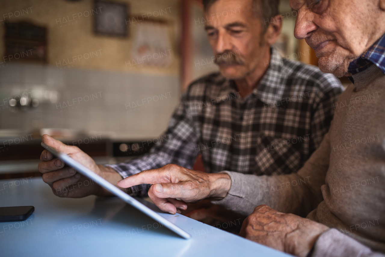 A portrait of man with elderly father sitting at the table indoors at home, using tablet.