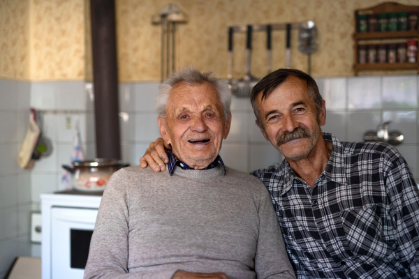 A portrait of man with elderly father sitting at the table indoors at home, looking at camera.