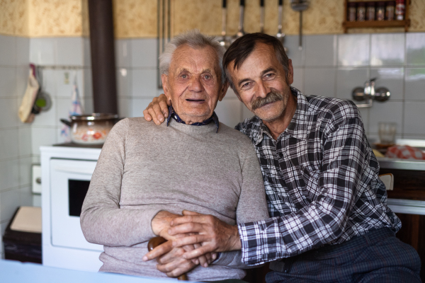 A portrait of man with elderly father sitting at the table indoors at home, looking at camera.