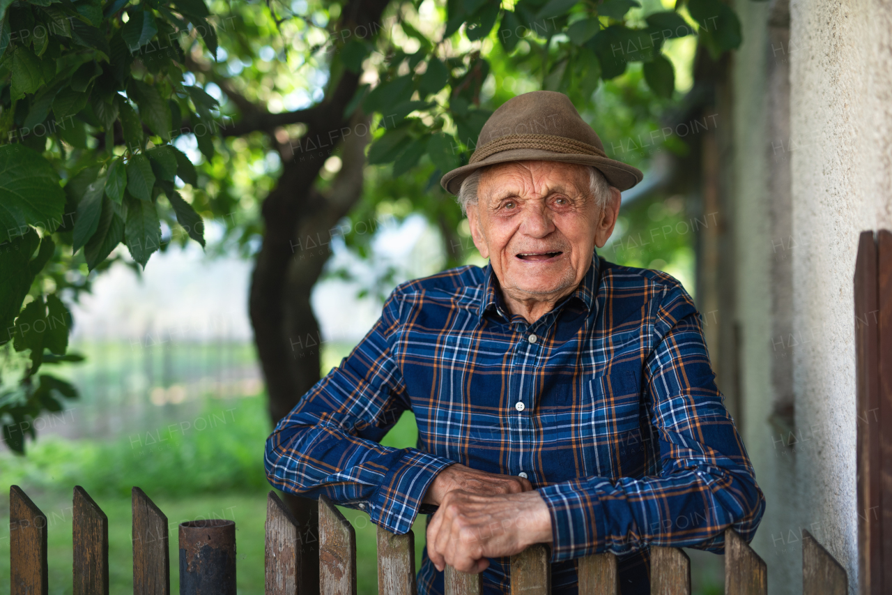 A portrait of elderly man standing outdoors in garden, leaning on wooden fence and looking at camera.