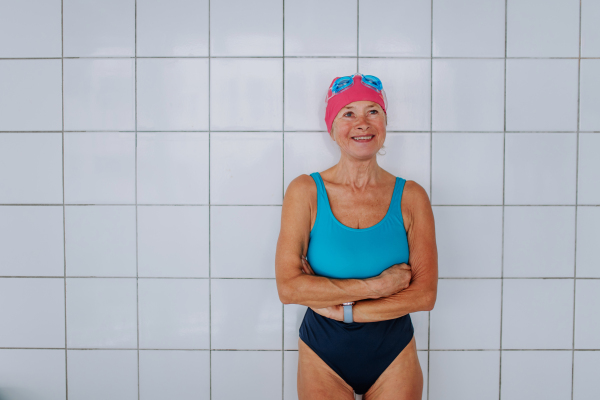 An active senior swimmer woman standing against white tiles wall indoors in swimming pool.