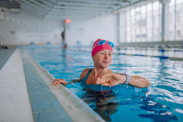 A senior woman looking at smartwatch when swimming in indoors swimming pool.