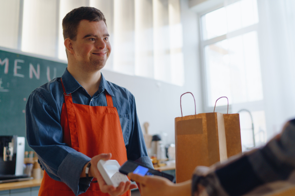 A cheerful young Down Syndrome waiter taking contactless smartphone payment from costumer in take away restaurant.