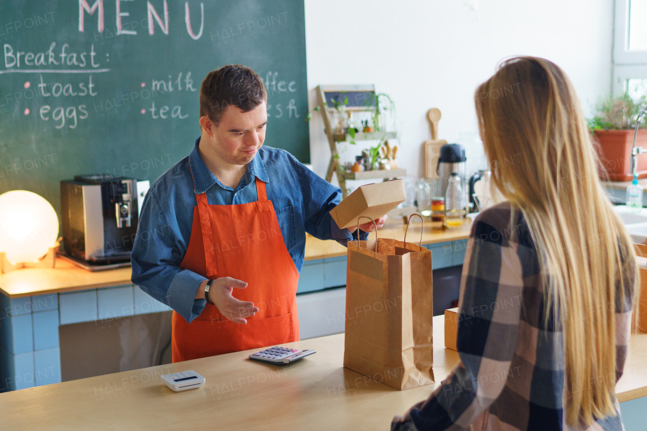 A cheerful young Down Syndrome waiter serving customer in take away restaurant, social inclusion concept.