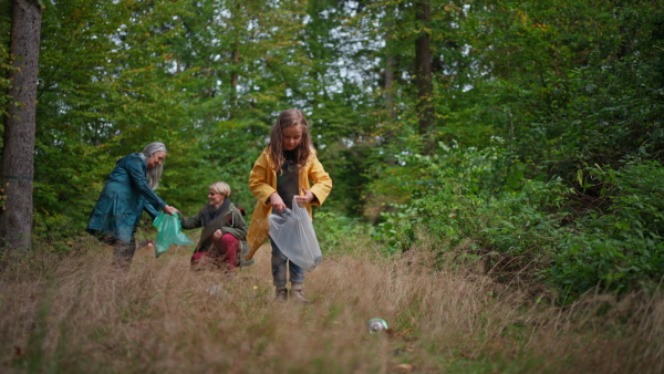 A small girl with mother and grandmother picking up waste outoors in forest.