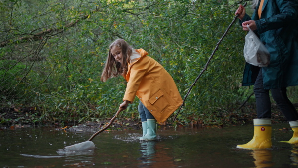 A small girl with grandmother picking up waste from little lake outoors in forest.