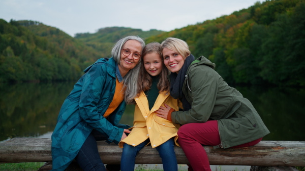 A small girl with mother and grandmother sitting on bench and looking at camera outoors by lake.