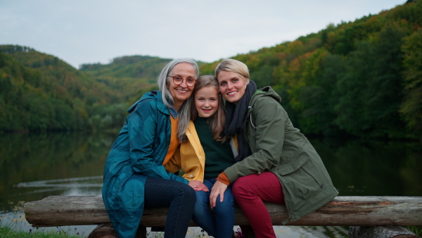 A small girl with mother and grandmother sitting on bench and looking at camera outoors by lake.