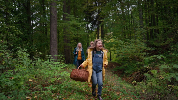 A happy little girl with basket running during walk with mother and grandmother outdoors in forest