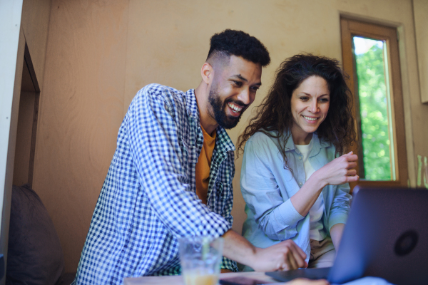 A happy couple with laptop sitting, resting and talking indoors in a tree house, remote office concept.