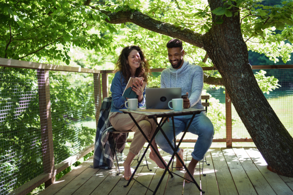 A happy couple with laptop resting outdoors in a tree house, weekend away and remote office concept.