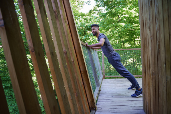 A high angle view of happy young man doing exercise outdoors on terrace of tree house, weekend away and digital detox concept.