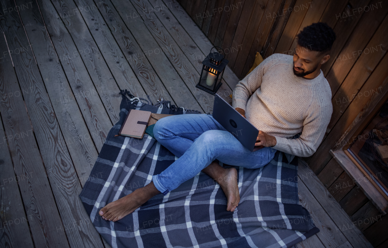 A happy young man with laptop resting outdoors in a tree house, weekend away and remote office concept. Working late at night. High angle shot.