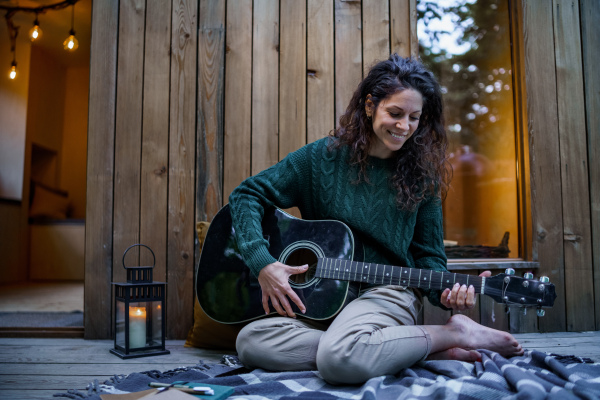 A happy mid adult woman playing acoustic guitar outdoors in a tree house, weekend away and digital detox concept. Female musican spending time alone in a cottage, composing songs on a wooden terrace.