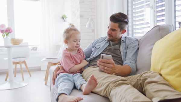 A young father sitting on sofa, holding his little daughter and using smartphone indoors at home