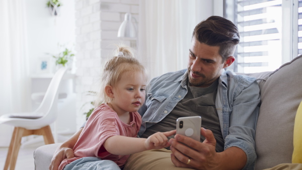 A young father sitting on sofa, holding his little daughter and using smartphone indoors at home