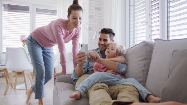 A young father sitting on sofa, holding his little daughter and using smartphone indoors at home