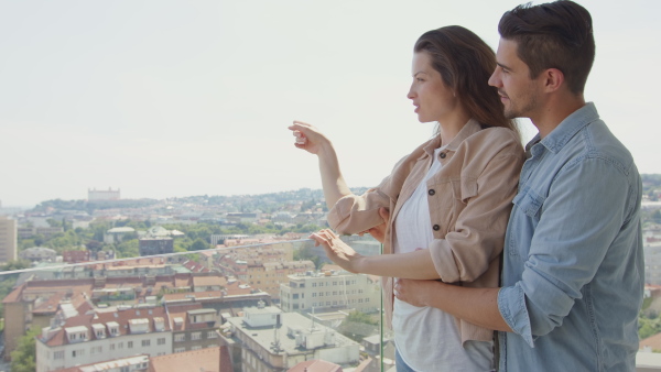 A side view of happy young couple hugging and looking at view on city from balcony.