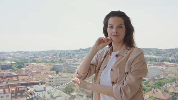 A portrait of young caucasian woman standing outdoors on balcony with urban view and looking at camera.