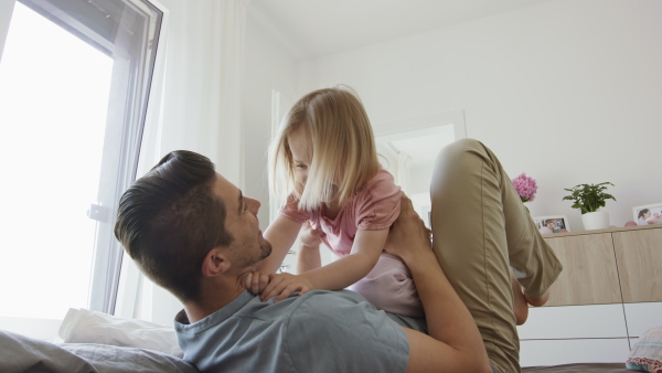 A happy young man lying on bed, holding his little daughter and having fun indoors at home.