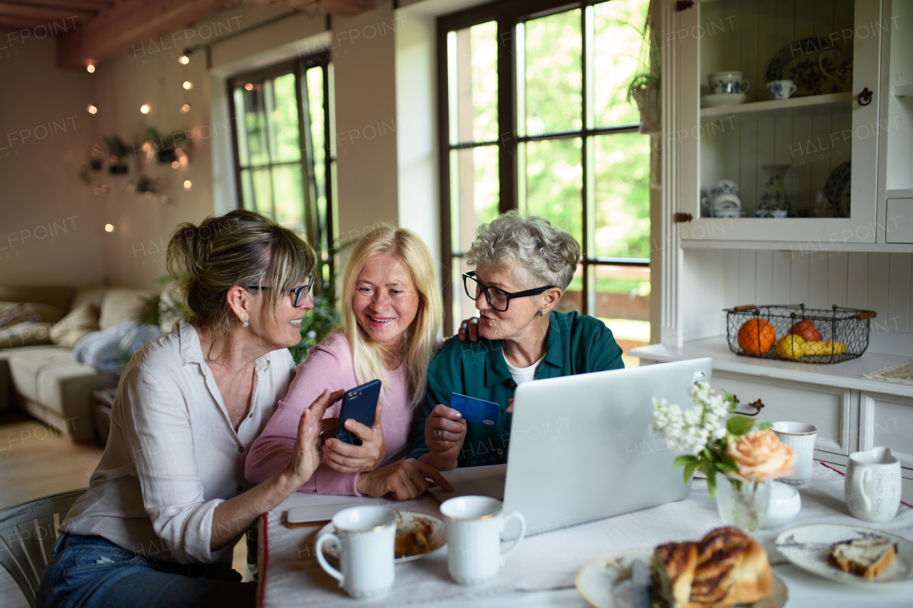 Happy senior friends using a laptop and smartphone indoors at home, coffee time.