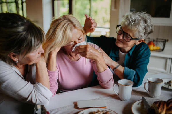 Female senior friends consloing a sad friend indoors at home.