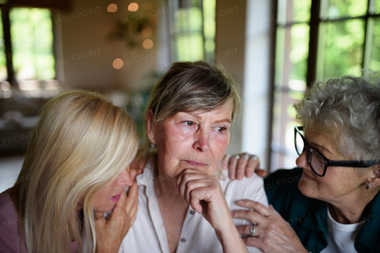 Female senior friends consloing a sad friend indoors at home.