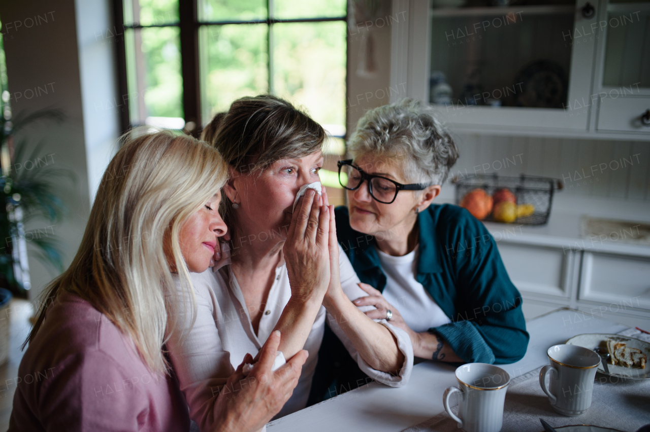 Female senior friends consloing a sad friend indoors at home.