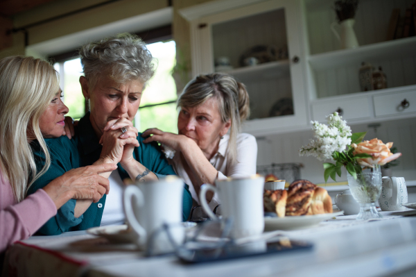 Female senior friends consloing a sad friend indoors at home.