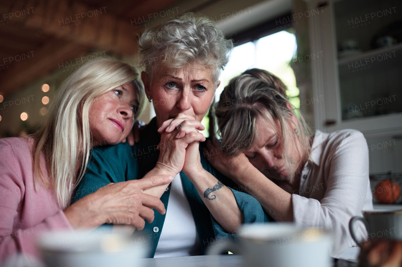 Female senior friends consloing a sad friend indoors at home.