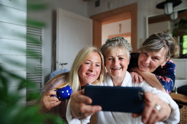 Happy senior women friends in bathrobes taking a selfie indoors at home, selfcare concept.