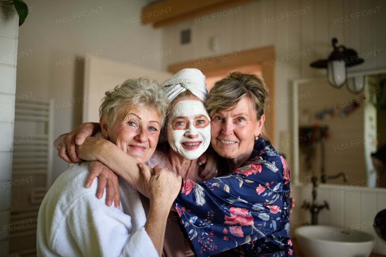 Happy senior women friends in bathrobes having fun indoors at home, looking at a camera.