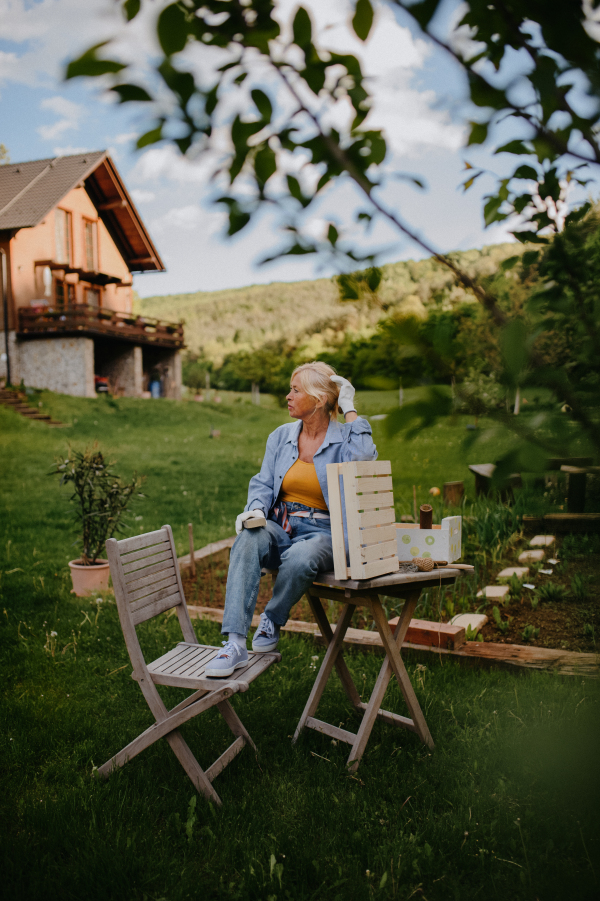 A happy senior woman renovating wooden crate outdoors in garden.