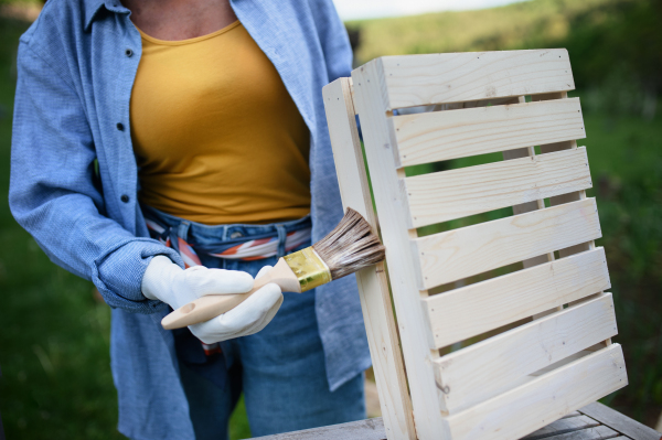 Close up of a senior woman hand with paintbrush impregnating wooden crate outdoors in garden.