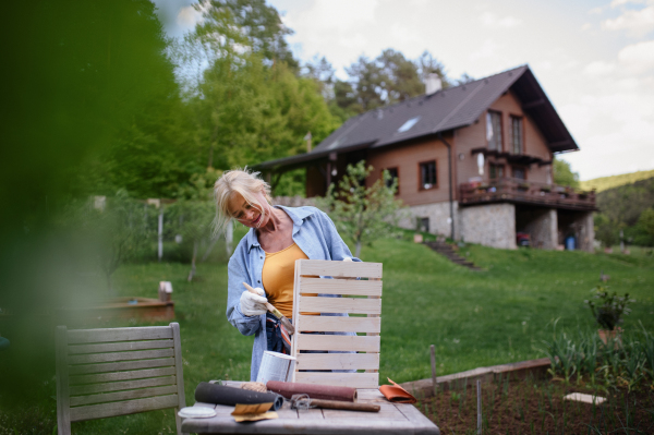 A senior woman with paintbrush impregnating wooden crate outdoors in garden.