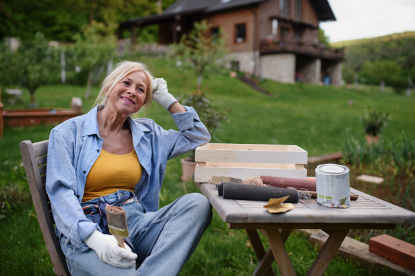 A happy senior woman sitting and resting while doing paint craft outdoors in garden.