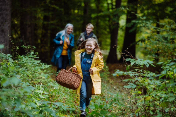 A happy little girl with basket running during walk with mother and grandmother outdoors in forest